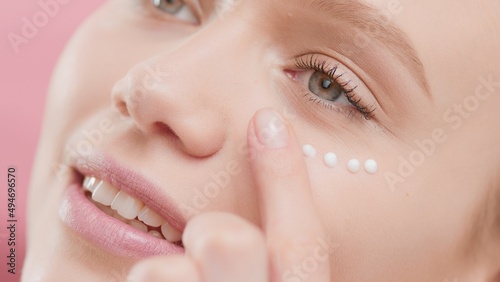 Extreme close-up shot of good-looking Caucasian woman who applies cream under her eye against pink background | Eye cream application and face care commercial concept