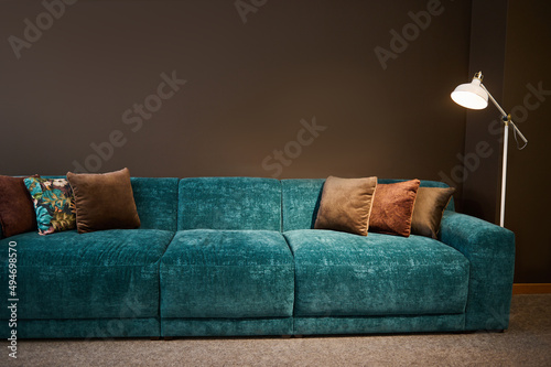 Exhibition of modern stylish upholstered furniture in the showroom of a furniture store. Focus on a turquoise soft velour setee and brown pillows lit by a lamp against a brown wall background photo