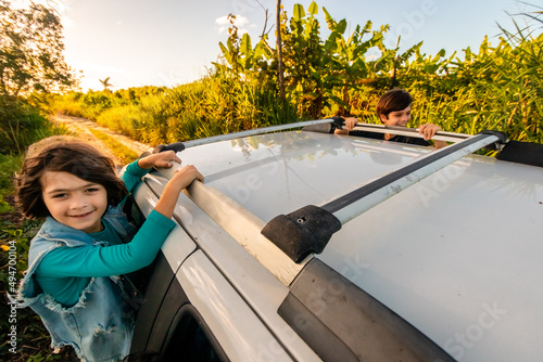Crianças sorrindo sentadas na janela de um carro numa estrada de terra. photo