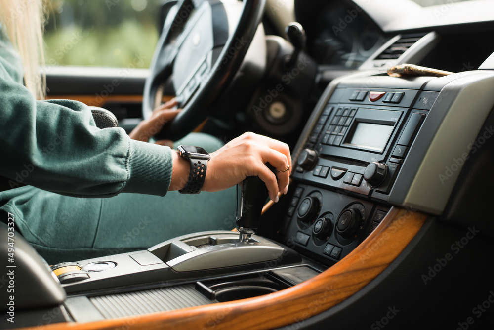 Caucasian female driver driving car, side view. Close-up of woman's hand holding an automatic transmission lever inside vehicle. Selective focus on female hand with watch