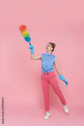 full length of tattooed young woman in blue rubber gloves holding dust brush on pink.