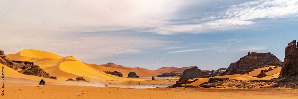 Fototapeta premium Panoramic view of Sahara Desert sand dune and rocky mountain off road nature.Tadrart Rouge, Djanet, Illizi. Erg dusty road. Orange colored sandstones and white sand powder in the air. Blue cloudy sky.