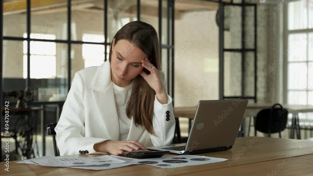 Stylish young female accountant looking through finances, using a laptop and a calculator, experiencing stress due to the fact that the numbers do not add up in the report.