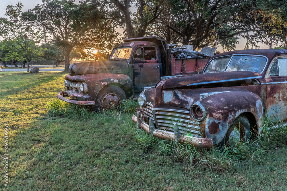 A pair of antique vehicles, a truck and a sedan, rusting away on a grassy field with oak trees and the sun rising in the background, Kendalia, Texas A pair of antique vehicles, a truck and a sedan, rusting away on a grassy field with oak trees and the sun rising in the background, Kendalia, Texas