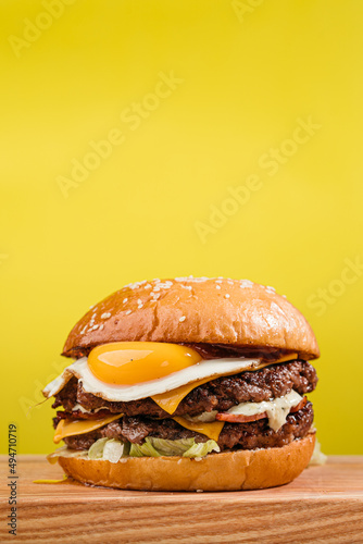 Double patty burger with marbled beef, bacon, cheddar cheese, fried egg, tomatoes, onions, cream sauce and ketchup. Wooden table, yellow background