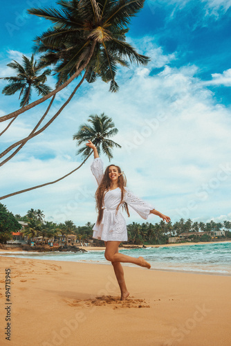party mood. young brunette girl in white summer dress is standing in jump with hands to the side and smiling happy on the beach coast background near palms. travel lifestyle concept, free space