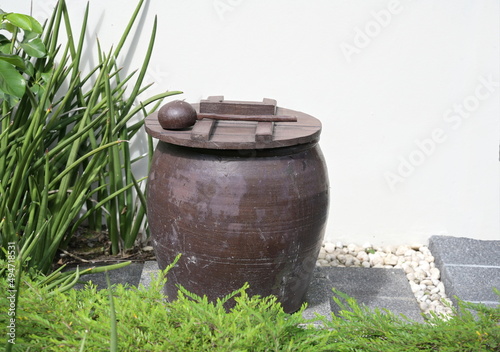 The old water jarwas placed in the garden beside the white wall of the sunlit house. A large clay pot with a wooden lid covered with a dipper or a coconut shell ladle is placed on top.
