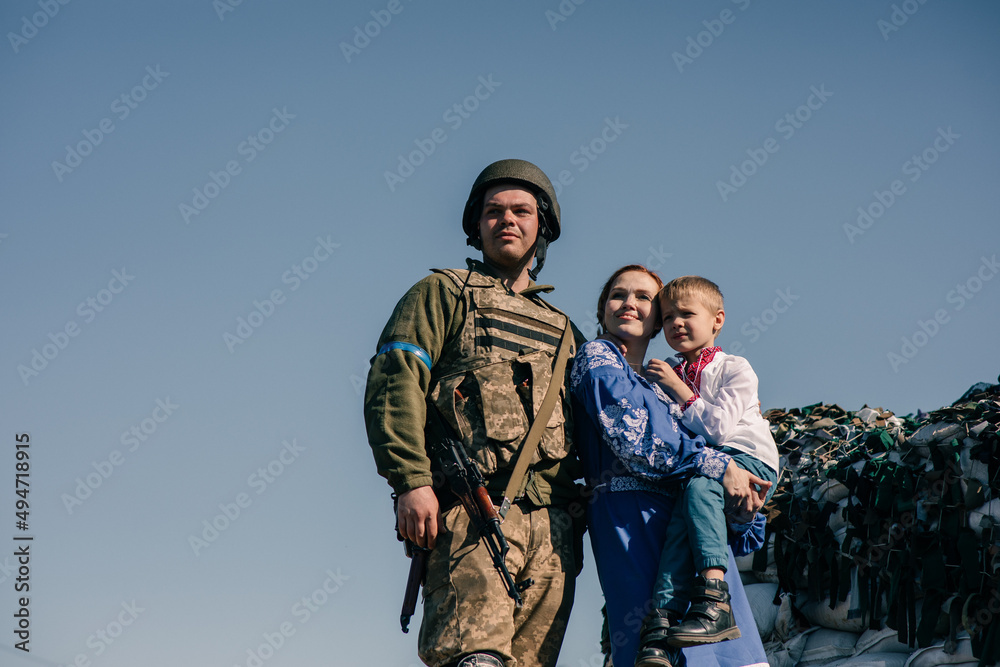 Woman with child boy on her hands stands near Ukrainian soldier on checkpoint against sandbags and sky background.
