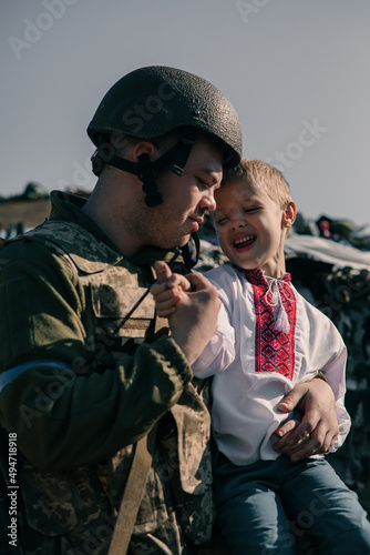Ukrainian soldier embraces child boy in embroidered shirt sitting near him on checkpoint.