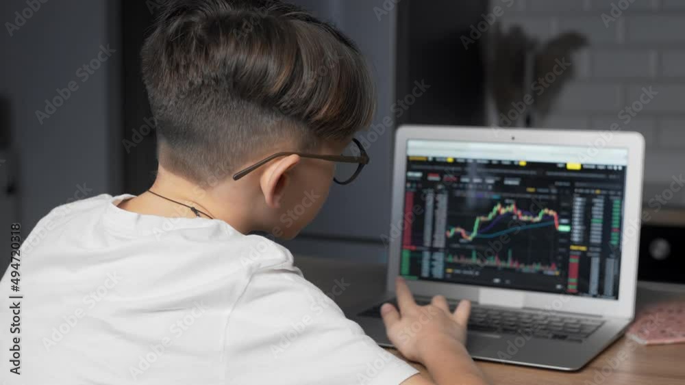 The boy is sitting at the computer and looking at an Online Stock Market Chart Showing Bearish And Bullish Trends Of The Bitcoin Currency. In real time The boy is sitting at the computer and looking at an Online Stock Market Chart Showing Bearish And Bullish Trends Of The Bitcoin Currency. In real time