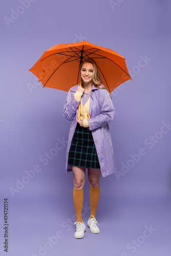 full length of happy young woman in trench coat standing under orange umbrella on purple.