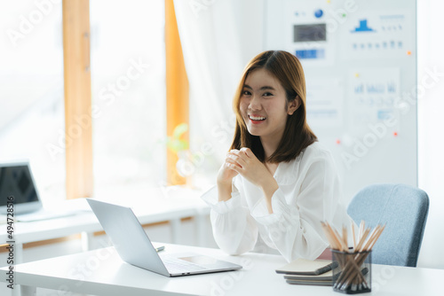 Portrait of Asian female executive sitting at office desk in front of laptop with financial business plan and smiling at camera at office © PaeGAG