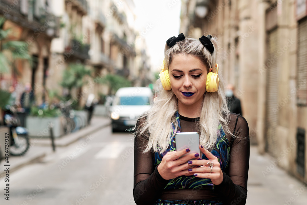 Obraz premium Young alternative woman using a mobile phone with headphones while walking down the street.