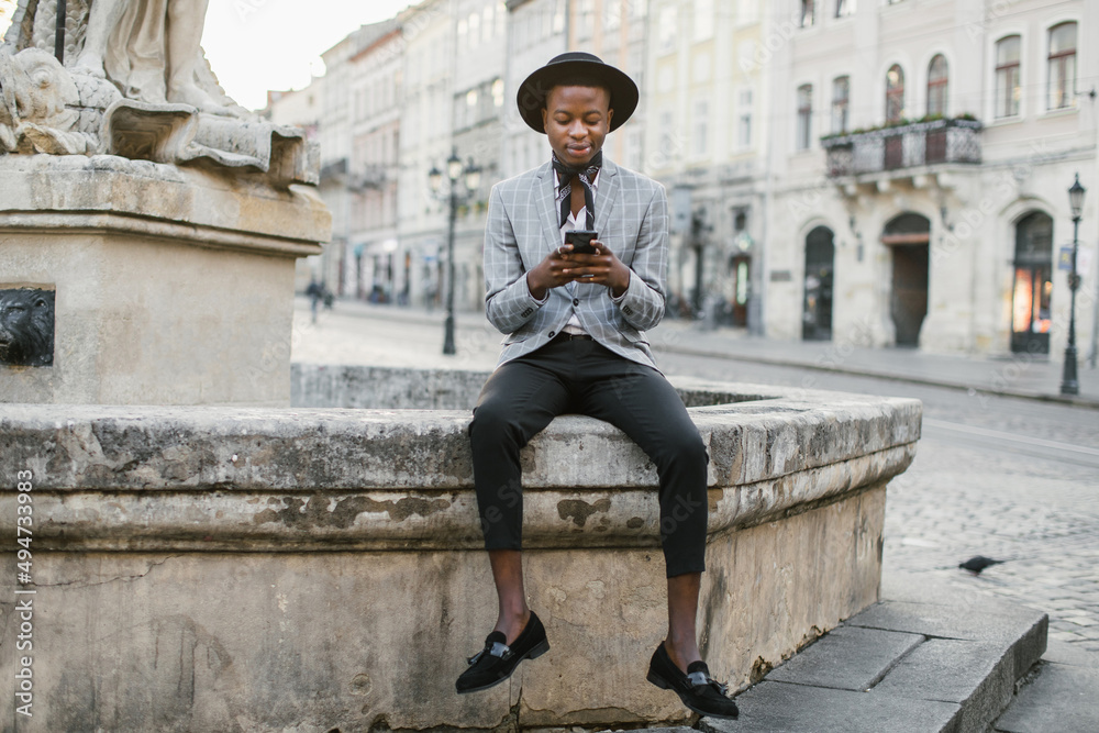 Happy african american man in suit and hat texting on modern smartphone while sitting outdoors. Blur background of street of old city. Happy african american man in suit and hat texting on modern smartphone while sitting outdoors. Blur background of street of old city.