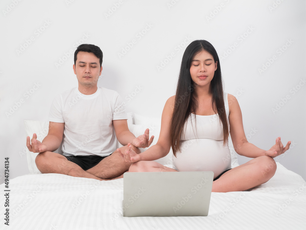 Asian couple, husband and pregnant wife sitting on the bed meditating with laptop infront of them.