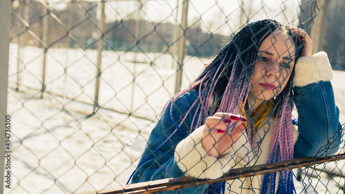 Stylish woman leaning on metal fence. Serious young female in warm clothes looking away while leaning on net fence on city street