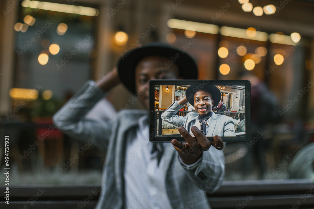 Close up of digital tablet with happy stylish african man on screen. Handsome young guy in hat and suit taking selfie while sitting on bench. Close up of digital tablet with happy stylish african man on screen. Handsome young guy in hat and suit taking selfie while sitting on bench.