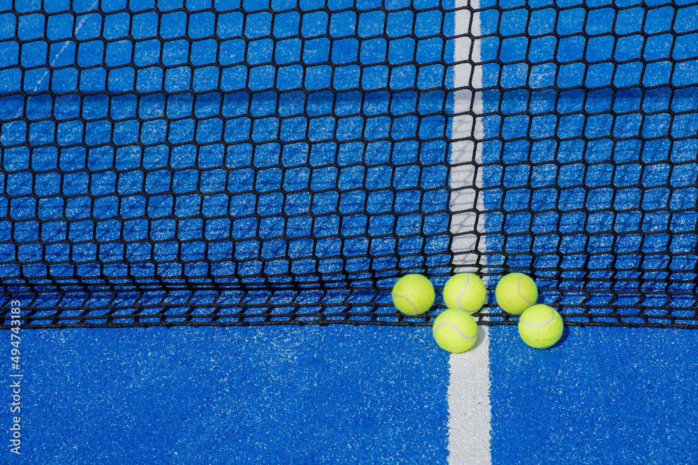 Five tennis balls by the net of a blue paddle tennis court, racket sports concept