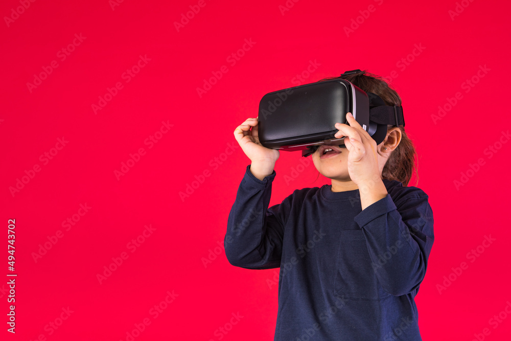 Side view of little girl wearing virtual reality goggles amazed and touching with her hands virtually, on red background. Virtual, augmented reality, technology, games and childhood concept. Side view of little girl wearing virtual reality goggles amazed and touching with her hands virtually, on red background. Virtual, augmented reality, technology, games and childhood concept.