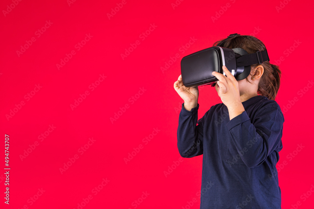 Side view of little girl wearing virtual reality goggles amazed and touching with her hands virtually, on red background. Virtual, augmented reality, technology, games and childhood concept. Side view of little girl wearing virtual reality goggles amazed and touching with her hands virtually, on red background. Virtual, augmented reality, technology, games and childhood concept.