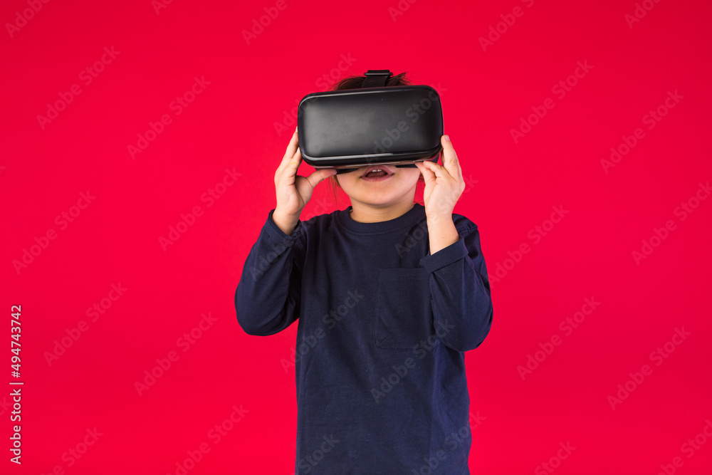 Front view of little girl wearing virtual reality glasses amazed and touching with her hands virtually, on red background. Virtual, augmented reality, technology, games and childhood concept. Front view of little girl wearing virtual reality glasses amazed and touching with her hands virtually, on red background. Virtual, augmented reality, technology, games and childhood concept.