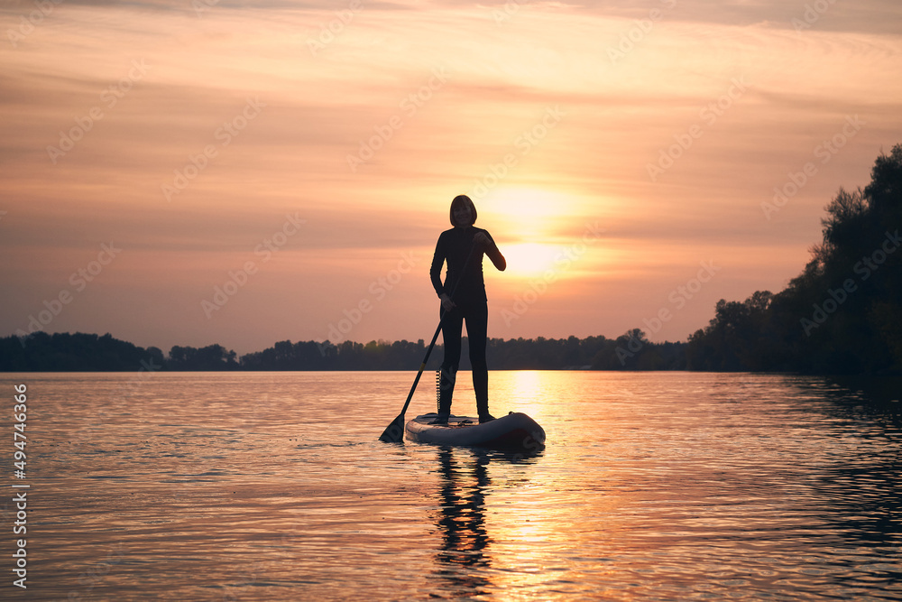 Silhouette of woman paddle on stand up paddle boarding (SUP) on quiet autumn river at twilight. Calm river surface after sunset. Sports and meditation on the water