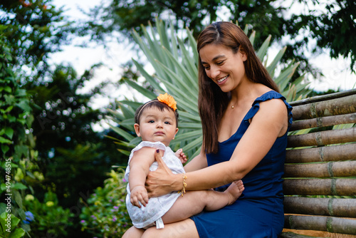 latina mother smiling looking at her baby sitting on her lap, sitting on a bench in nature