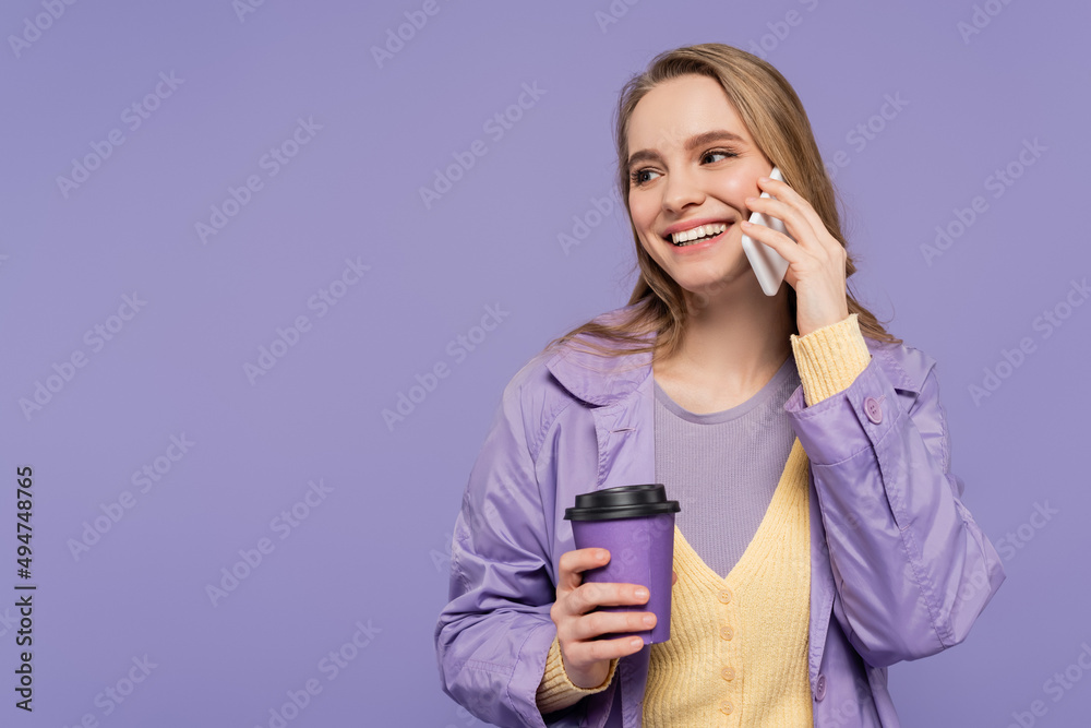 smiling young woman in trench coat talking on smartphone and holding paper cup isolated on purple. smiling young woman in trench coat talking on smartphone and holding paper cup isolated on purple.