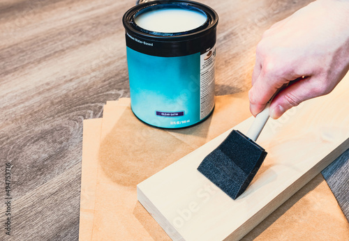 Hand using foam brush testing a clear matte  coating protection paint on sample of birch wood before marking built-in furniture for new house with wooden floor background. DIY home improvement concept