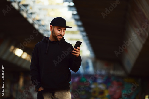 Leinwand Poster Young bearded man with a black hat  and black hoodie in the city using smartphon