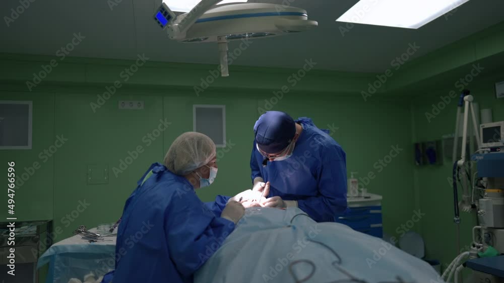 Concentrated surgeon and assistant doing operation on patient face in surgery room indoors. Professional confident Caucasian man and woman performing reconstructive surgery in medical clinic Concentrated surgeon and assistant doing operation on patient face in surgery room indoors. Professional confident Caucasian man and woman performing reconstructive surgery in medical clinic