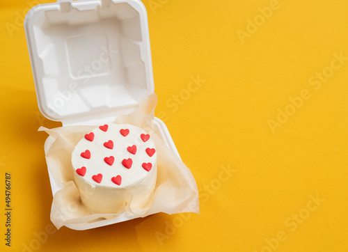 Bento cake in the form of circle with small red hearts in eco packaging. Picture for websites about desserts, food, confectionery. Yellow background with space for text. Selective focus.