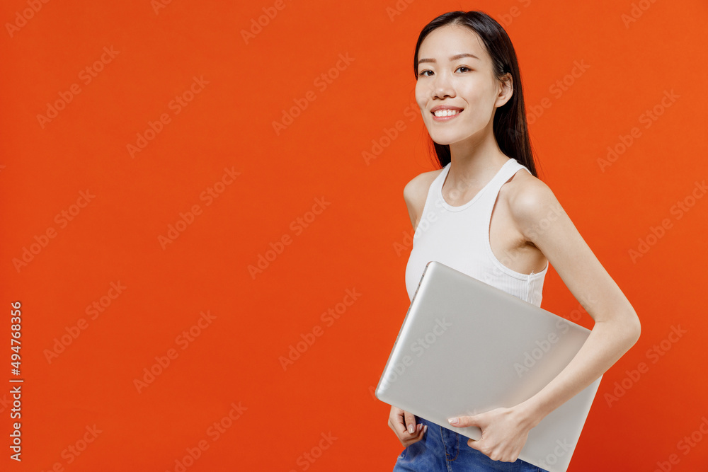 Side profile view fun smiling young woman of Asian ethnicity 20s years old in white tank top hold use work on laptop pc computer stepping strolling isolated on plain orange background studio portrait. Side profile view fun smiling young woman of Asian ethnicity 20s years old in white tank top hold use work on laptop pc computer stepping strolling isolated on plain orange background studio portrait.