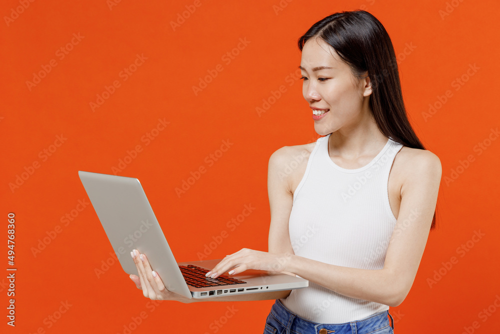 Smiling vivid young woman of Asian ethnicity 20s years old in white tank top hold use work on laptop pc computer typing browsing chatting send sms isolated on plain orange background studio portrait. Smiling vivid young woman of Asian ethnicity 20s years old in white tank top hold use work on laptop pc computer typing browsing chatting send sms isolated on plain orange background studio portrait.