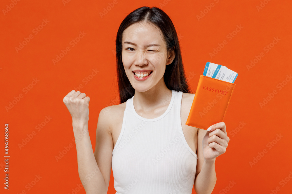 Young woman of Asian ethnicity 20s years old in white tank top hold passport boarding tickets doing winner gesture clenching fists say yes blinking isolated on plain orange background studio portrait. Young woman of Asian ethnicity 20s years old in white tank top hold passport boarding tickets doing winner gesture clenching fists say yes blinking isolated on plain orange background studio portrait.