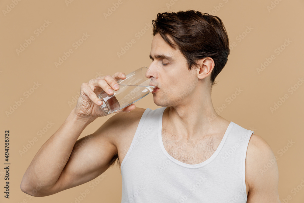 Attractive european young man 20s perfect skin wear undershirt hold glass of water drink isolated on pastel pastel beige background studio portrait. Skin care healthcare cosmetic procedures concept. Attractive european young man 20s perfect skin wear undershirt hold glass of water drink isolated on pastel pastel beige background studio portrait. Skin care healthcare cosmetic procedures concept.