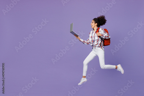 Full size fun young girl woman of African American ethnicity teen student in shirt hold backpack jump high hurry up run hold laptop pc computer isolated on plain purple background. Education concept.