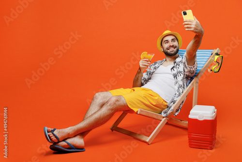 Canvas Print Young tourist man wear beach shirt hat lie on deckchair near fridge drink cocktail do selfie shot on mobile cell phone isolated on plain orange background