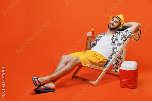 Fototapeta Young smiling happy fun cool tourist man in beach shirt hat hold beer bottle lie on deckchair near fridge isolated on plain orange background studio portrait