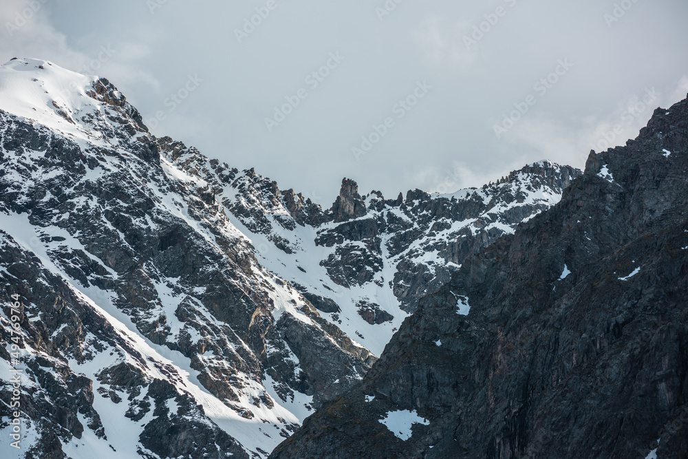 Dramatic landscape with sunlit high snowy mountain with peaked top in cloudy sky. White snow on black sharp rocks with golden shine. Awesome snow mountain peak with gold sunlight in changeable weather