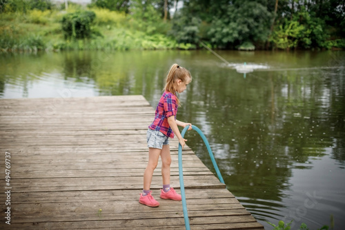 A blonde girl stands on a wooden pier on the river bank.