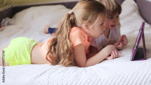 Two beautiful little sisters lying in the bed and look at the screen of a tablet, smart kids using smart technology