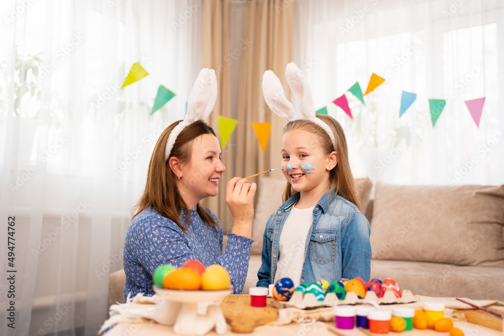 mother draws a rabbit with paints on her daughter's face to celebrate Easter.