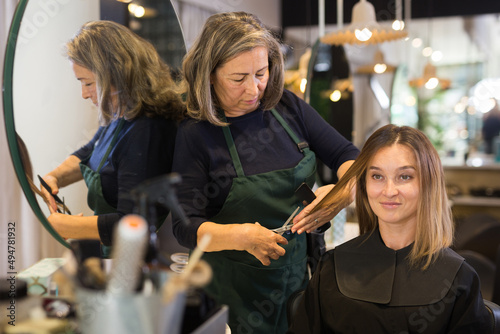 Attractive brown-haired young woman getting haircutting by professional elderly female hairdresser in modern hair salon.