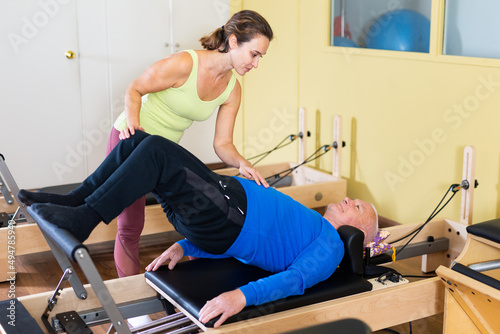 Personal female trainer controlling movements of mature man doing pilates on reformer in fitness studio. © JackF