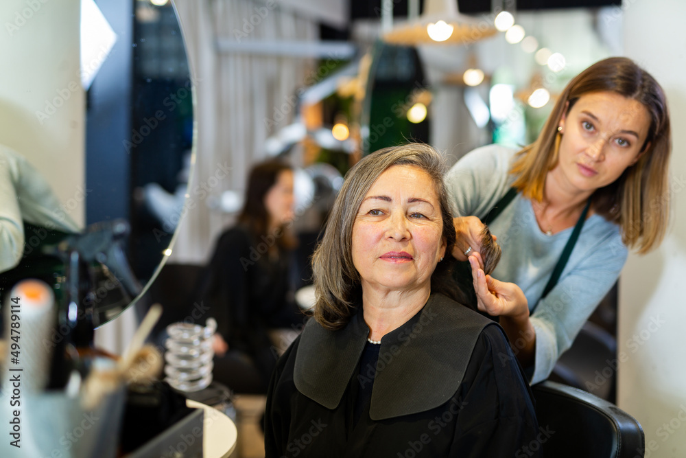 Positive elderly woman visiting professional hairdressing salon. Young female hairdresser examining hair of client.