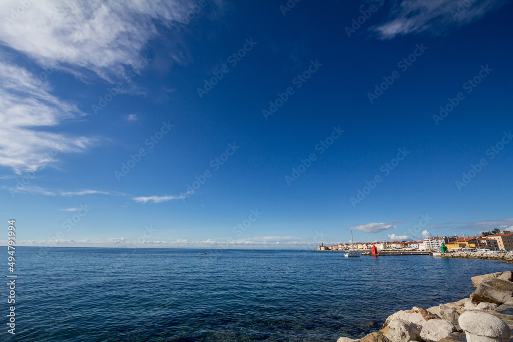 Panorama of  Piran, Slovenia, with Adriatic sea in front, with blue water and sky, on a wharf and quay, during a sunny summer afternoon. Piran, or Pirano, is a slovenian city on adriatic sea in istria
