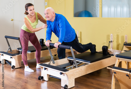 Focused positive senior man practicing pilates system on reformer to improve and maintain mobility under supervision of qualified female trainer