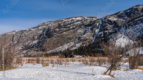 A picturesque mountain against a blue sky. The houses of the tourist base are visible at the foot. Dry grass and bare trees in a snowy valley. Altai