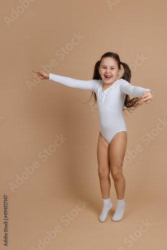 Portrait of young cute happy smiling girl with long dark hair in white training swimsuit and socks jumping on brown background, spreading hands, posing, standing on toes. Health, initial gymnastics. 